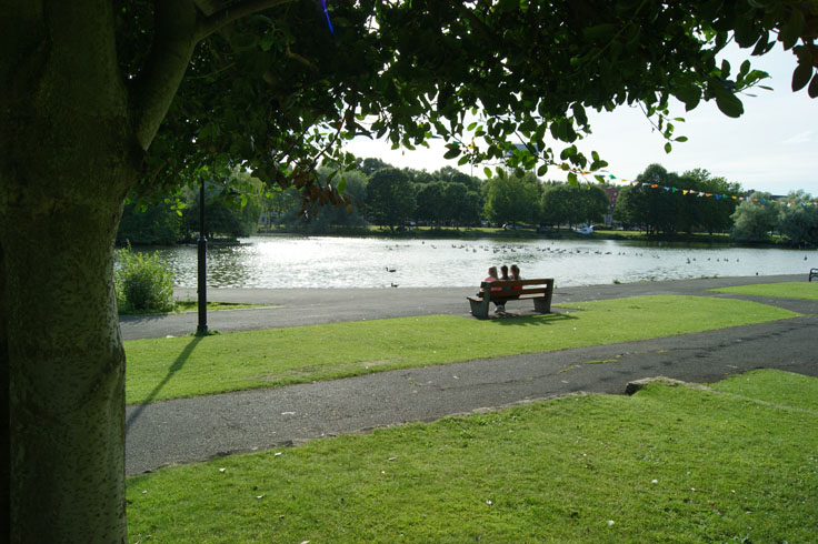 A group of people sat on a bench in front of a boating lake.